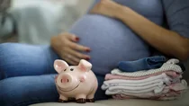 Neben einer schwangeren Frau stehen ein Sparschwein und ein Stapel mit Babykleidung.